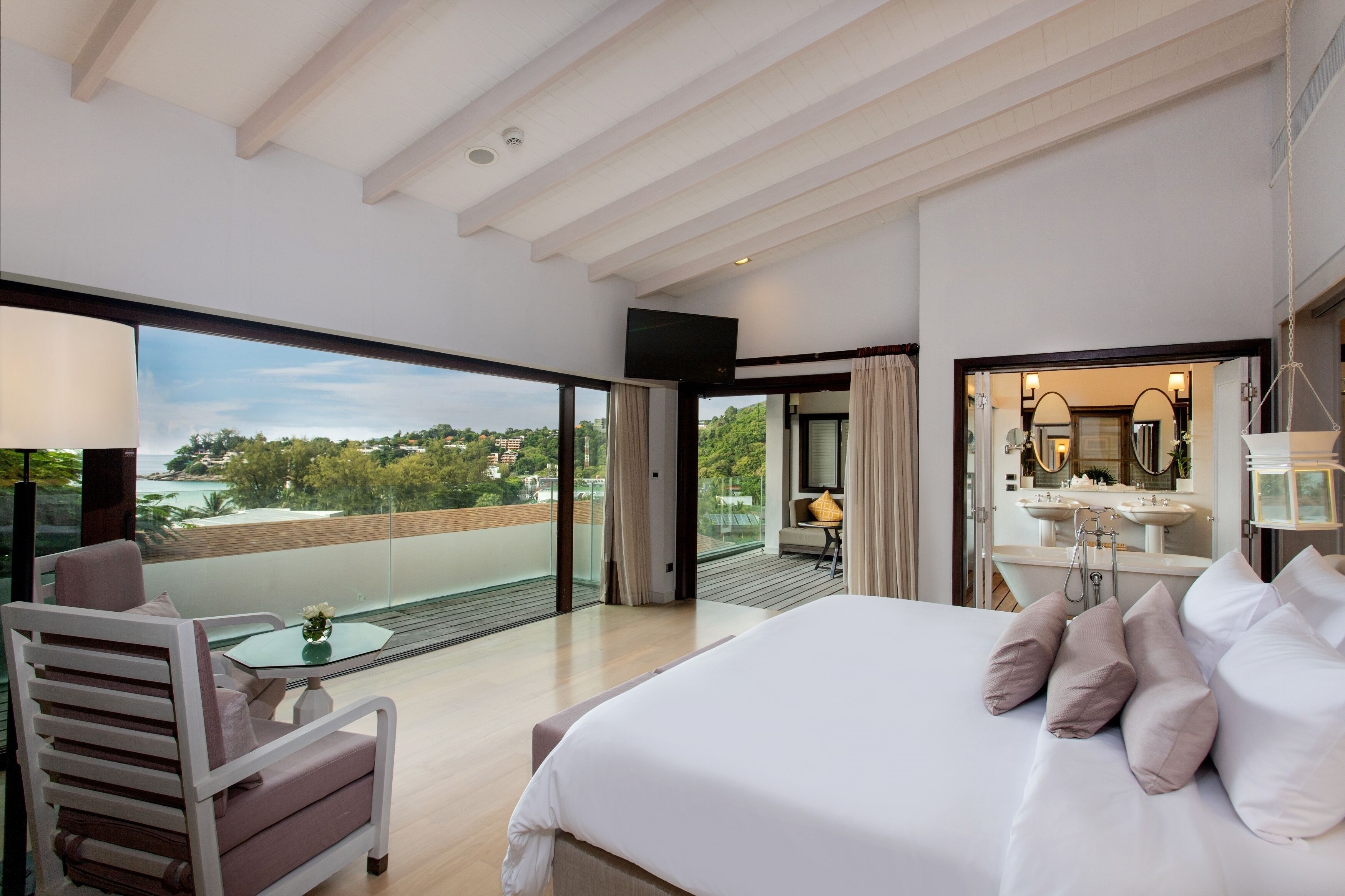 A modern hotel room with a large bed, a balcony with a view, a sitting area, and a bathroom with a large mirror.
