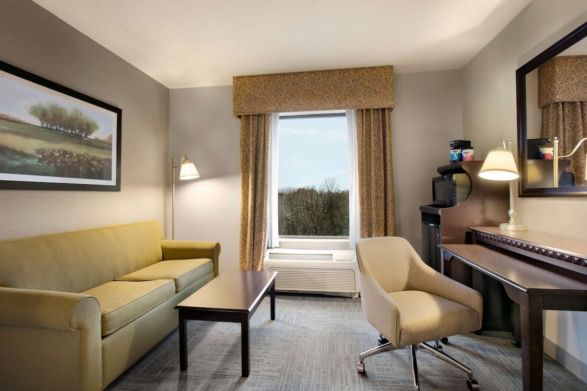A hotel room with a sofa, armchair, desk, and a large window with curtains.