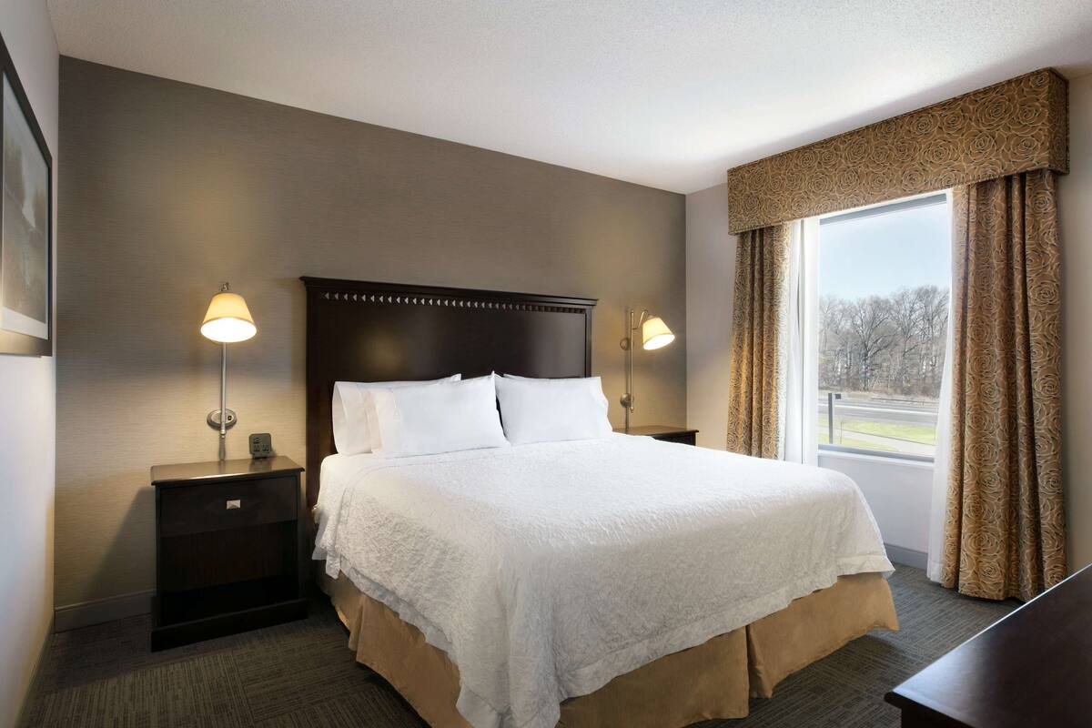 A neatly made bed with a white bedspread and pillows, two bedside tables with lamps, a large window with curtains, and a dark headboard.