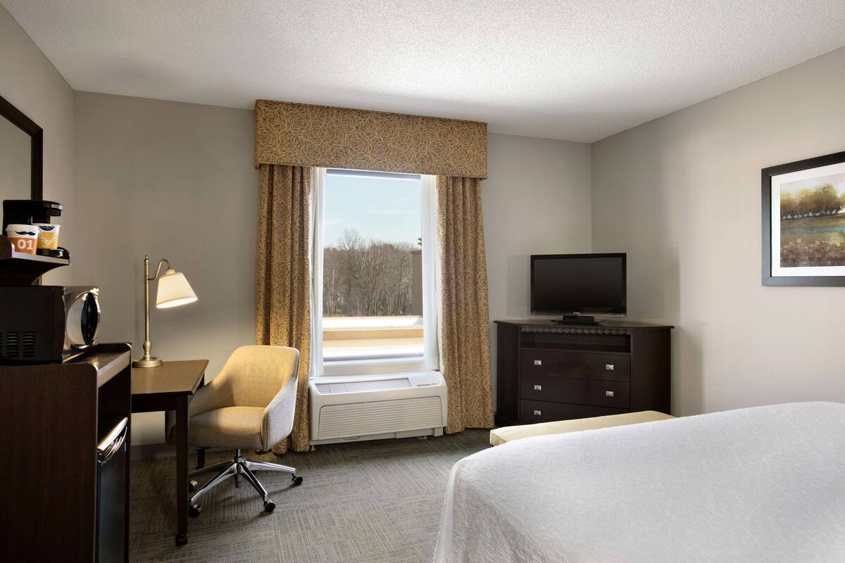 A hotel room with a bed, a desk with a lamp, a chair, a TV on a dresser, and a window with curtains.