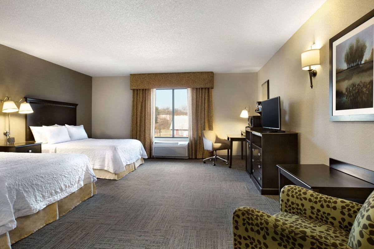 A hotel room with two beds, a desk, a chair, a TV, and a large window.