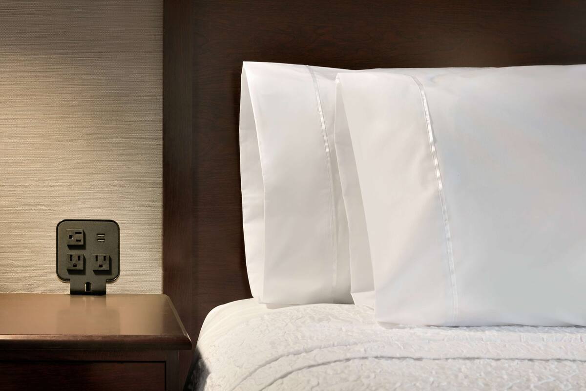 A neatly made bed with white linens and a wooden nightstand with a power outlet.