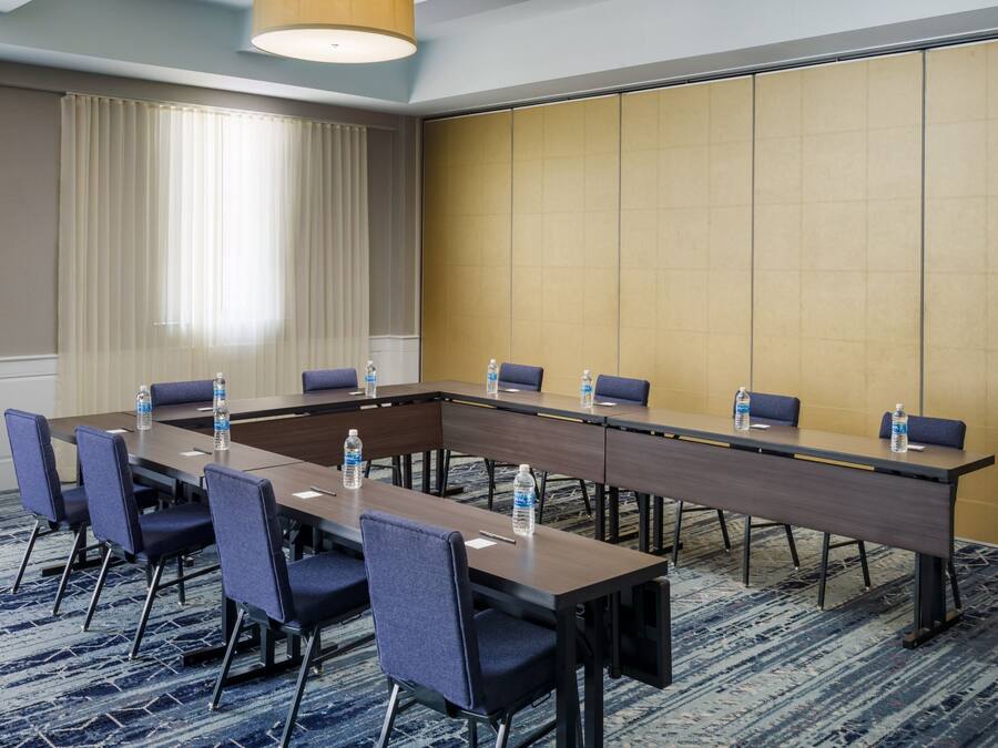 Un salón de conferencias con una larga mesa, sillas, botellas de agua y piso alfombrado.