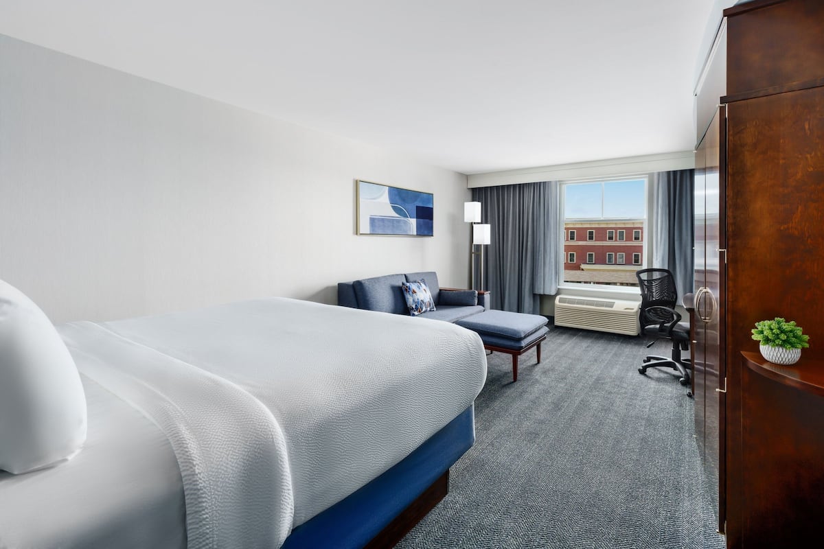 A hotel room with a large bed, a desk, a chair, and a view of a building through the window.