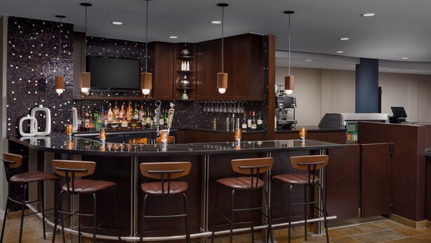 A bar area with a dark tiled backsplash, multiple stools, and a variety of bottles and glasses on the counter. - Courtyard by Marriott Keene Downtown (Keene)