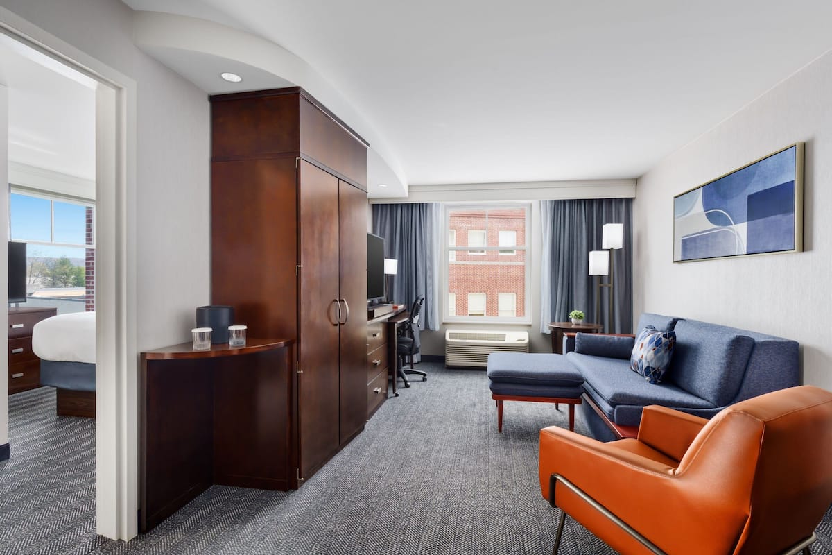 A hotel room with a large wardrobe, a sofa, an orange armchair, a desk with a chair, and a television.