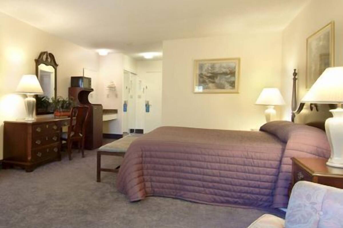 A hotel room with a bed, two bedside tables with lamps, a dresser, a chair, and a painting on the wall.