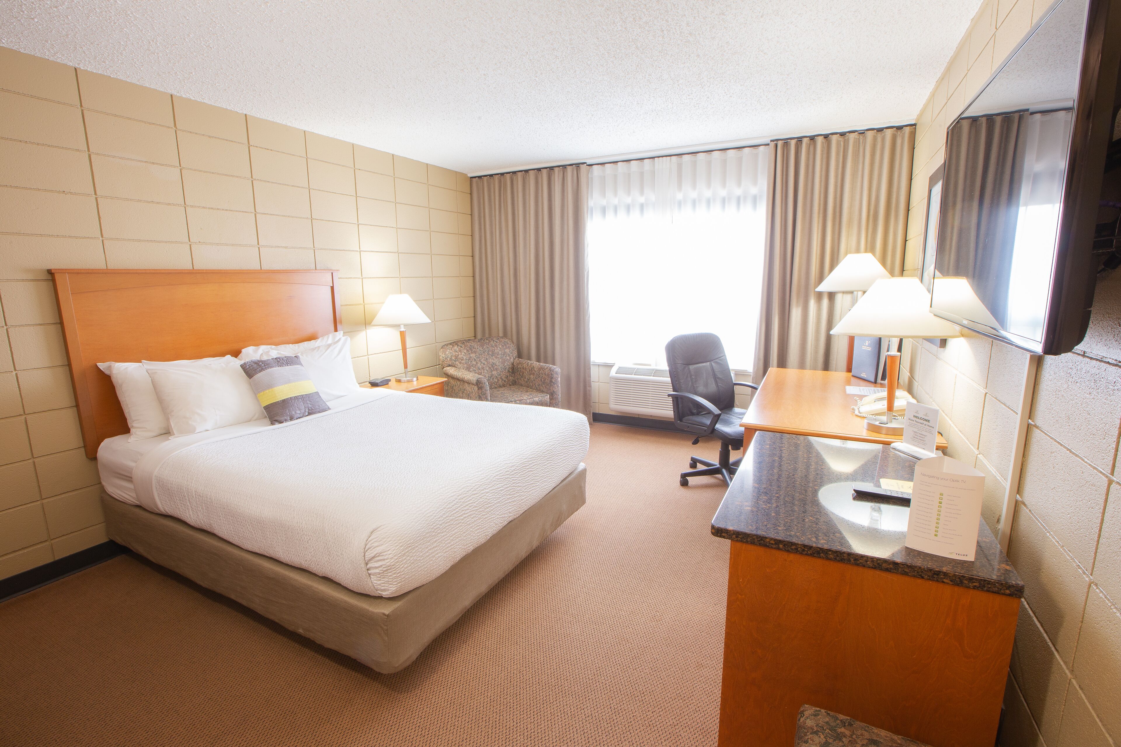 A hotel room with a large bed, a desk with a computer, a chair, a desk lamp, a mirror, and a window with curtains.