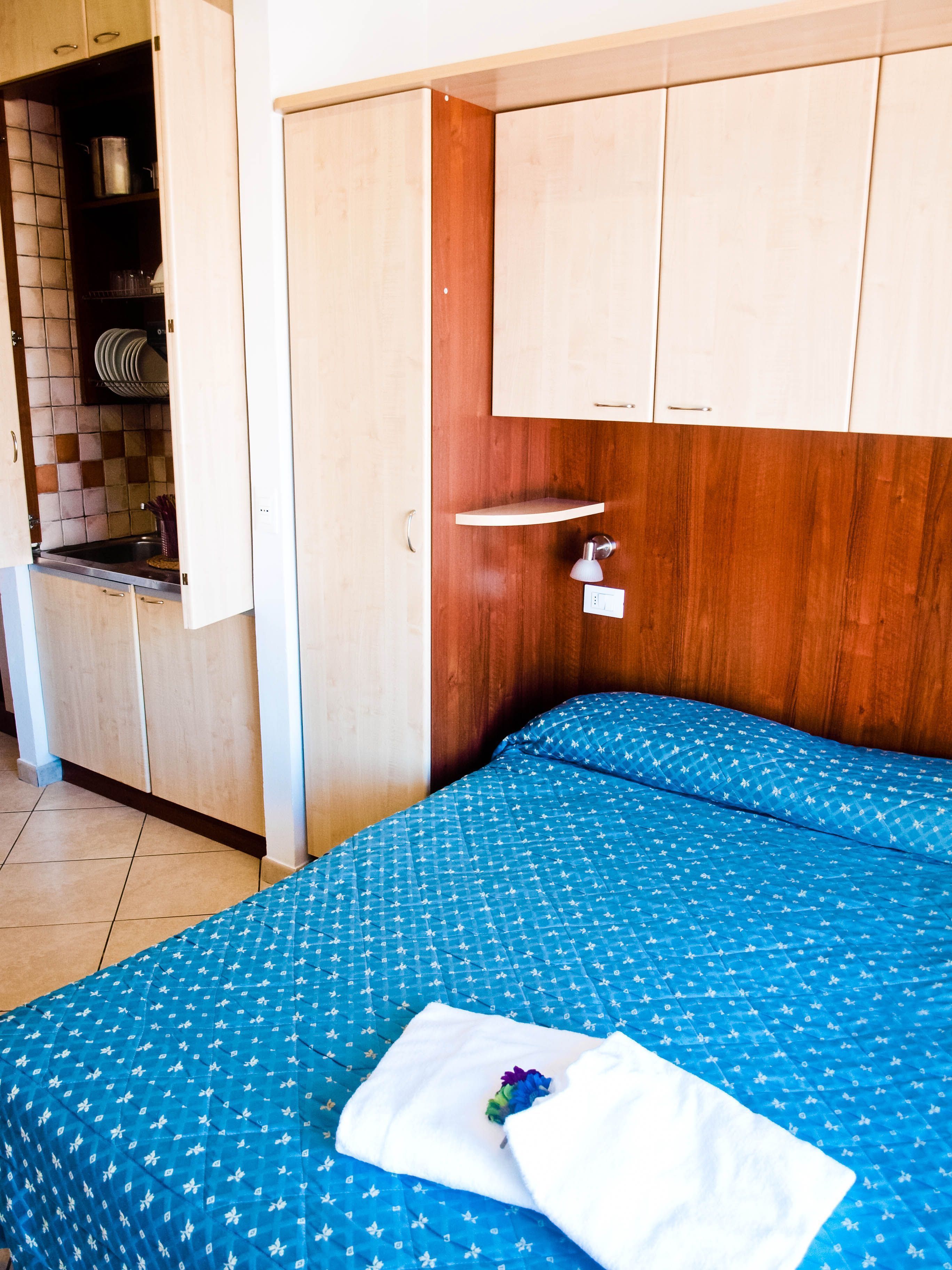 Une chambre à coucher comprenant un lit, une table de chevet, une armoire en bois et un coin cuisine visible à travers une porte ouverte.