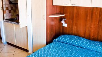 Quarto com cama, mesa de cabeceira, guarda-roupa de madeira e área de cozinha visível através de uma porta aberta.