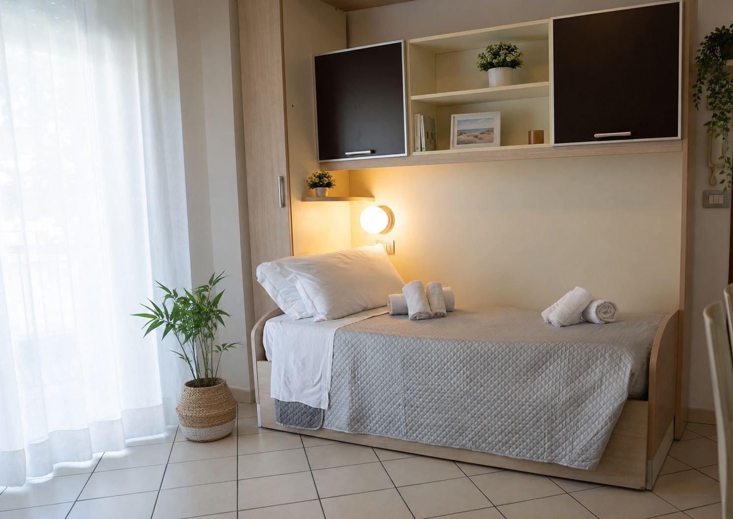 A neatly arranged bedroom with a bed, bedside lamps, a bedside plant, and a shelf with decorative items.