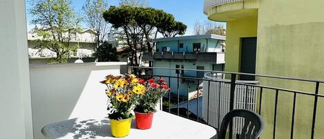 A balcony with a table and two chairs, potted plants, and a view of buildings and trees.