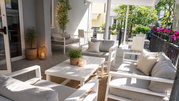 Zona de estar al aire libre con mobiliario blanco, incluyendo sofás y mesas, rodeada de plantas e iluminación decorativa.