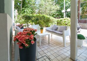 Un patio con un sofá blanco, una mesa pequeña y una planta en maceta con flores rosadas.