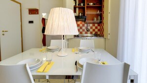 Un coin repas avec une table dressée pour deux personnes, une lampe et une cuisine en arrière-plan.