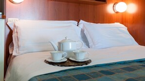 Un lit bien fait, avec du linge de lit blanc, une table de chevet sur laquelle se trouvent une théière blanche et des tasses, et deux lampes fixées au mur.