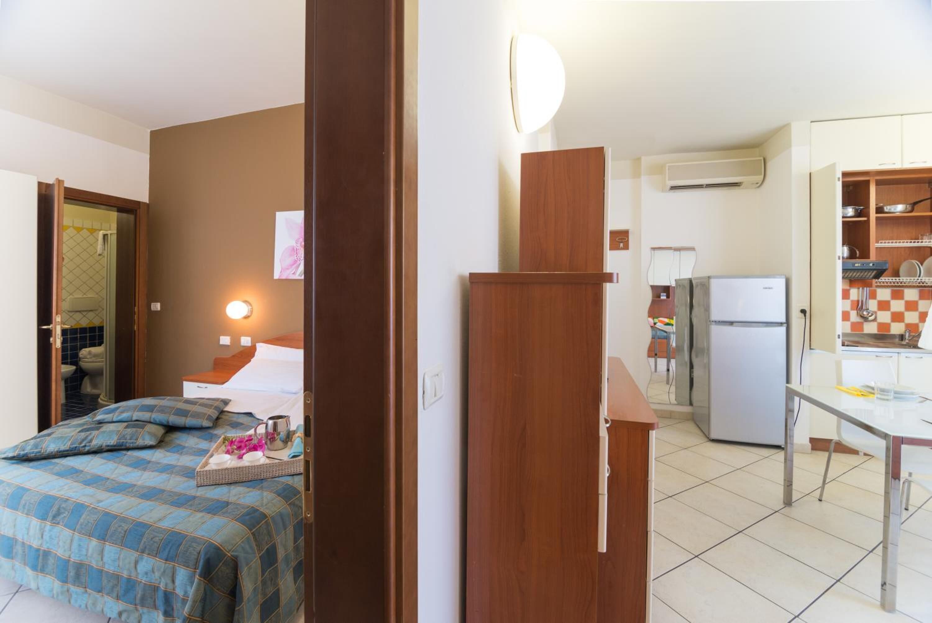 A hotel room with a bed, a bedside table, a nightstand, a mirror, a wall-mounted light, a wooden door, a kitchen area with a refrigerator, a sink, and a countertop.