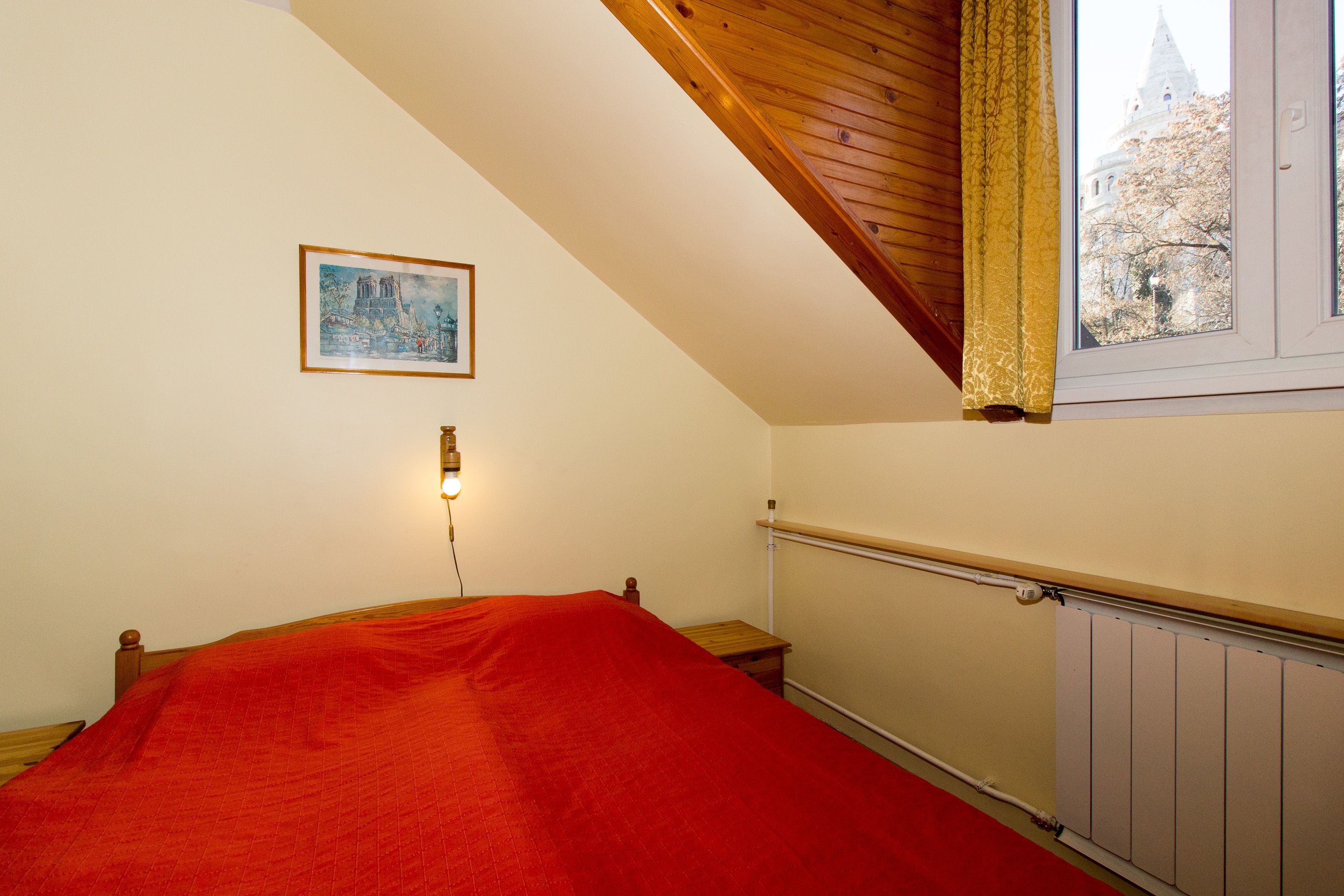 Et soveværelse med skråvæg, en seng med rødt tæppe, et natbord, en vægmonteret lampe, en radiator og et vindue med udsigt til en bygning.