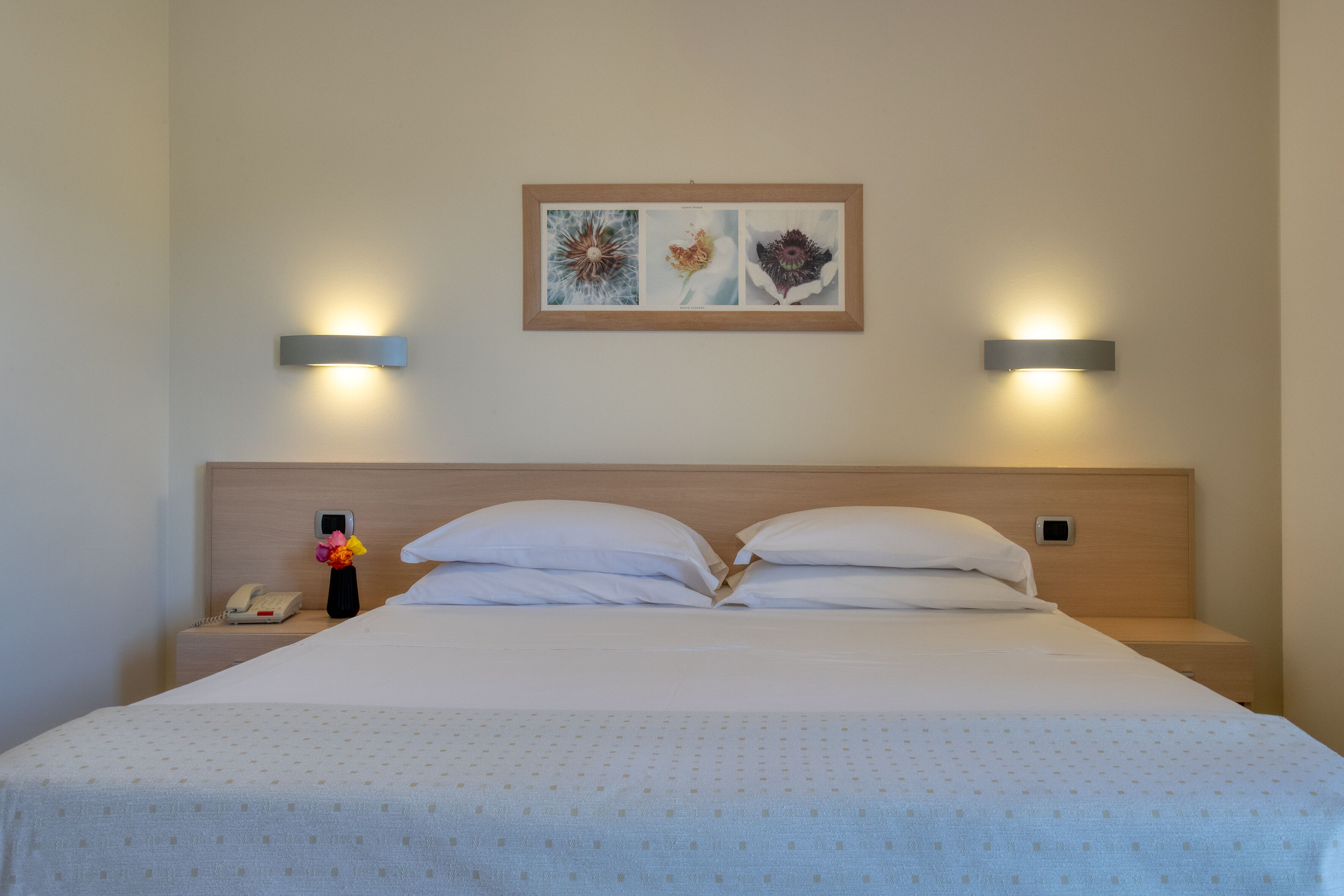 Un letto rifatto con cura, con una coperta a motivi, due lampade sui comodini, un comodino con un telefono e un vaso di fiori, e un quadro incorniciato sopra il letto.