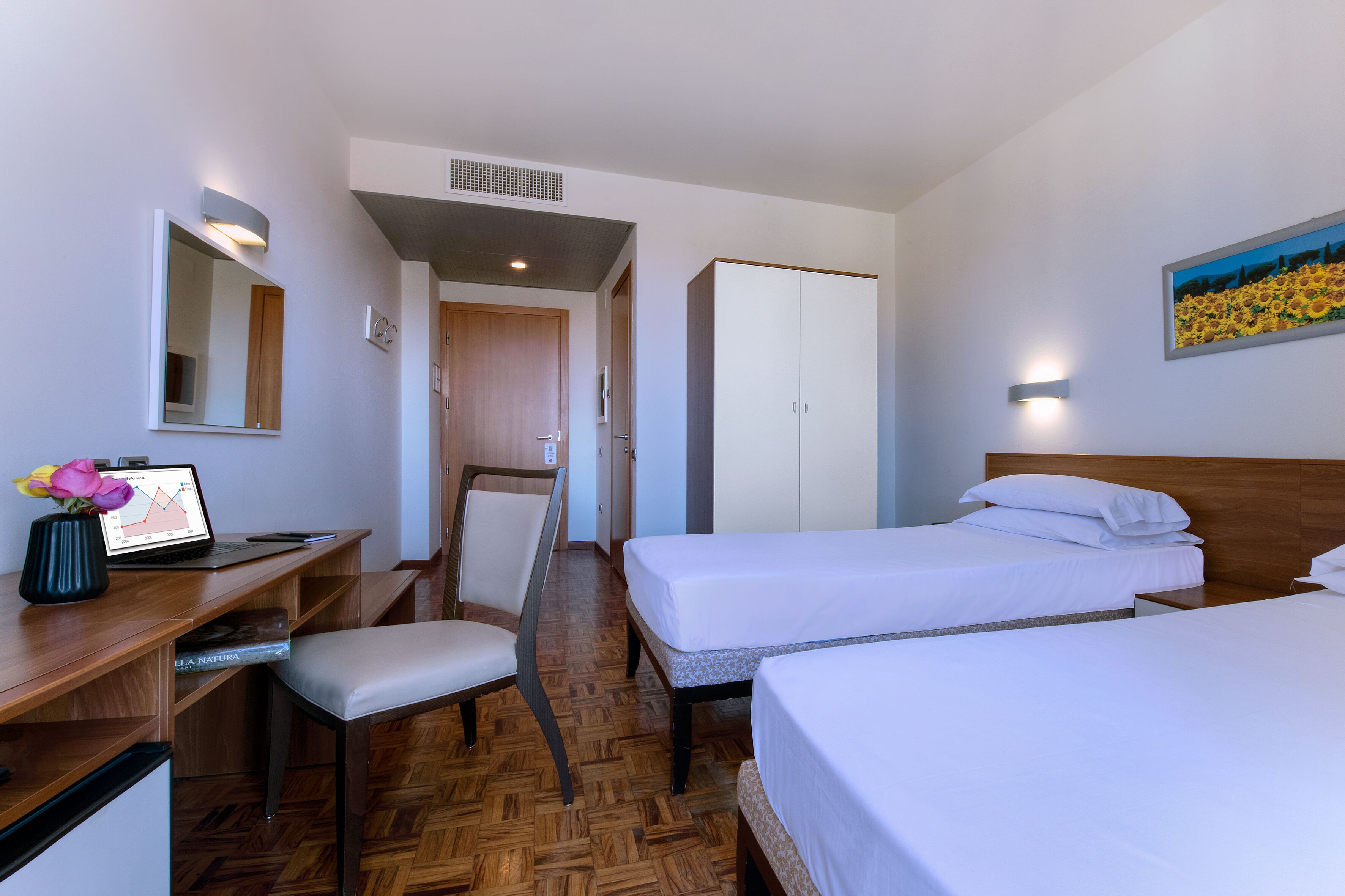 A hotel room with a wooden floor, a desk with a laptop, a chair, two beds, a wardrobe, and a painting on the wall.