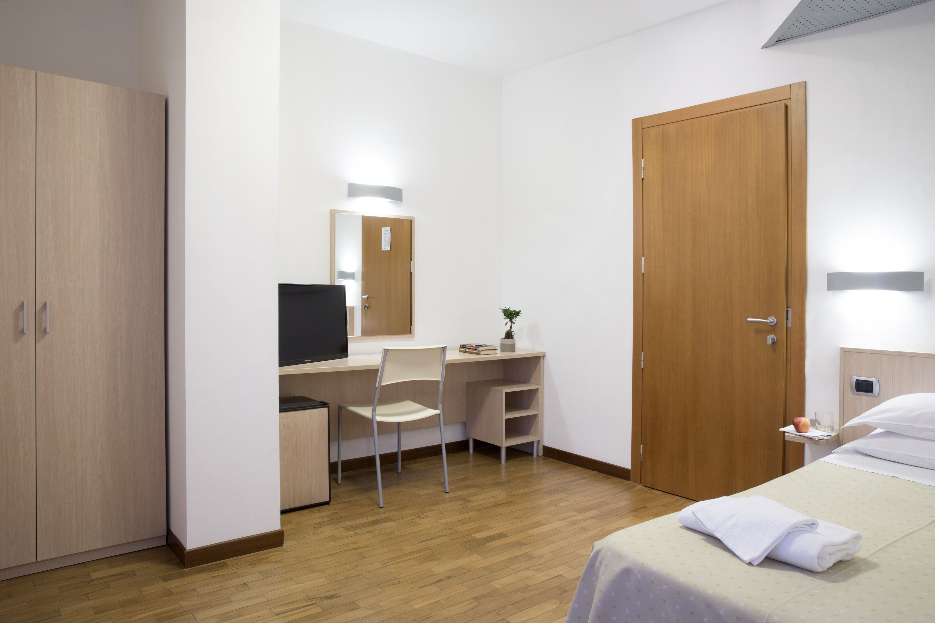 Une chambre d’hôtel avec une armoire en bois, un bureau avec une chaise, un lit avec une serviette blanche et une porte en bois.
