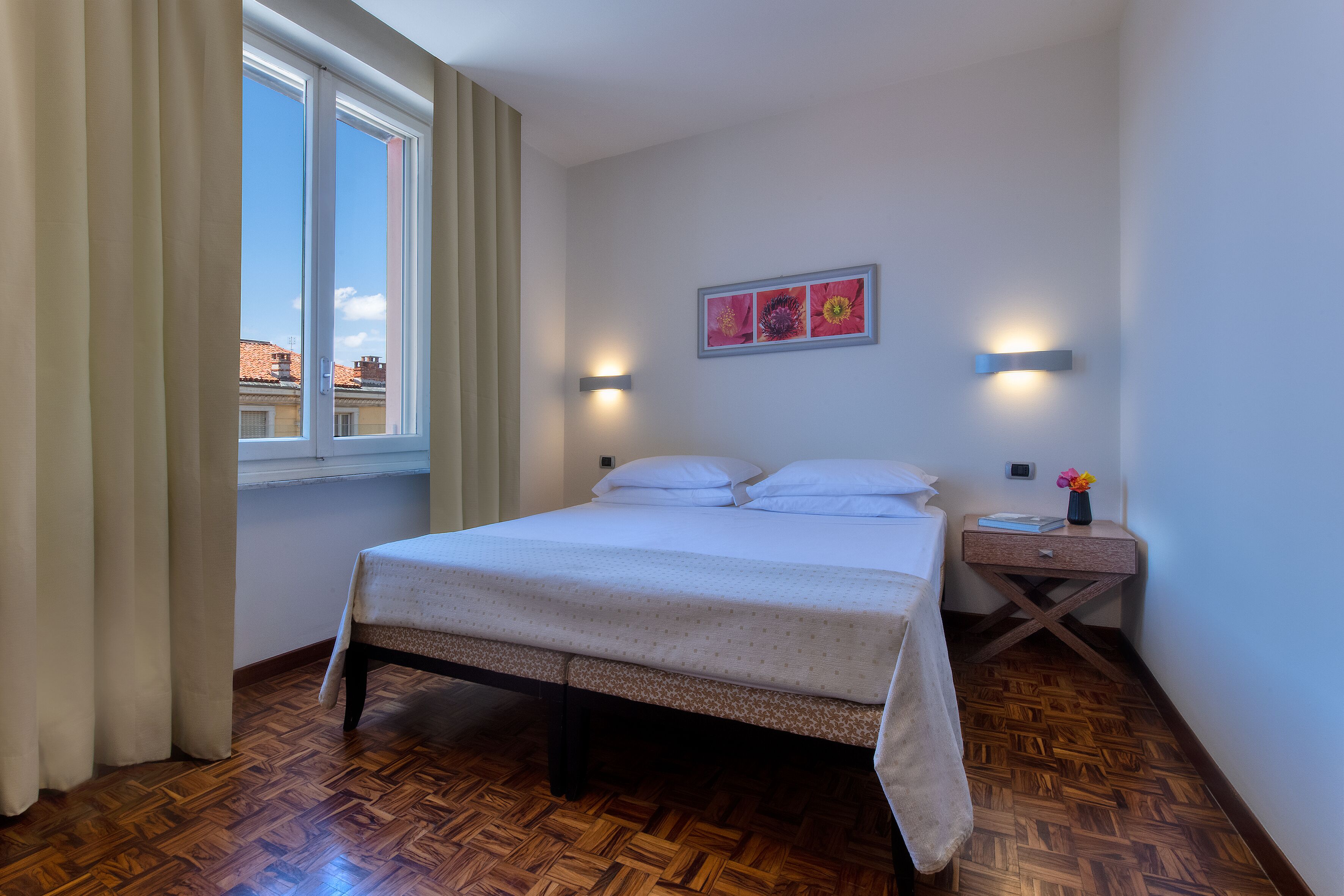 Un dormitorio con piso de parquet de madera, una cama con colcha blanca, una mesita de noche con un florero y una ventana con cortinas.