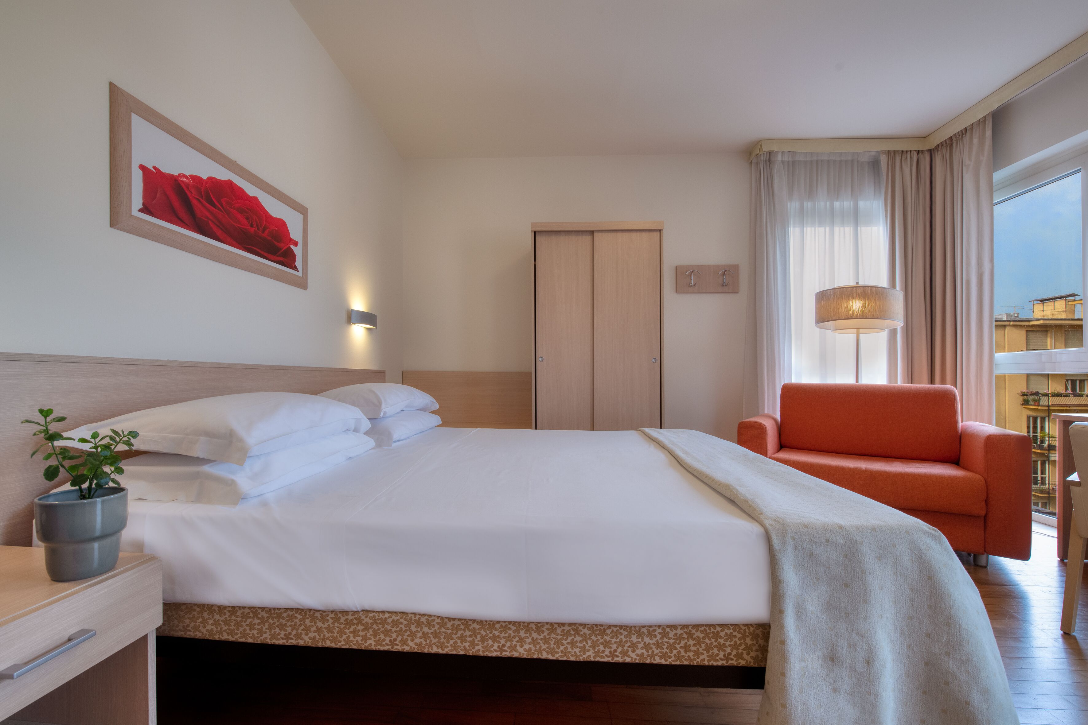 A hotel room with a bed, a chair, a desk, a potted plant, and a window with curtains.