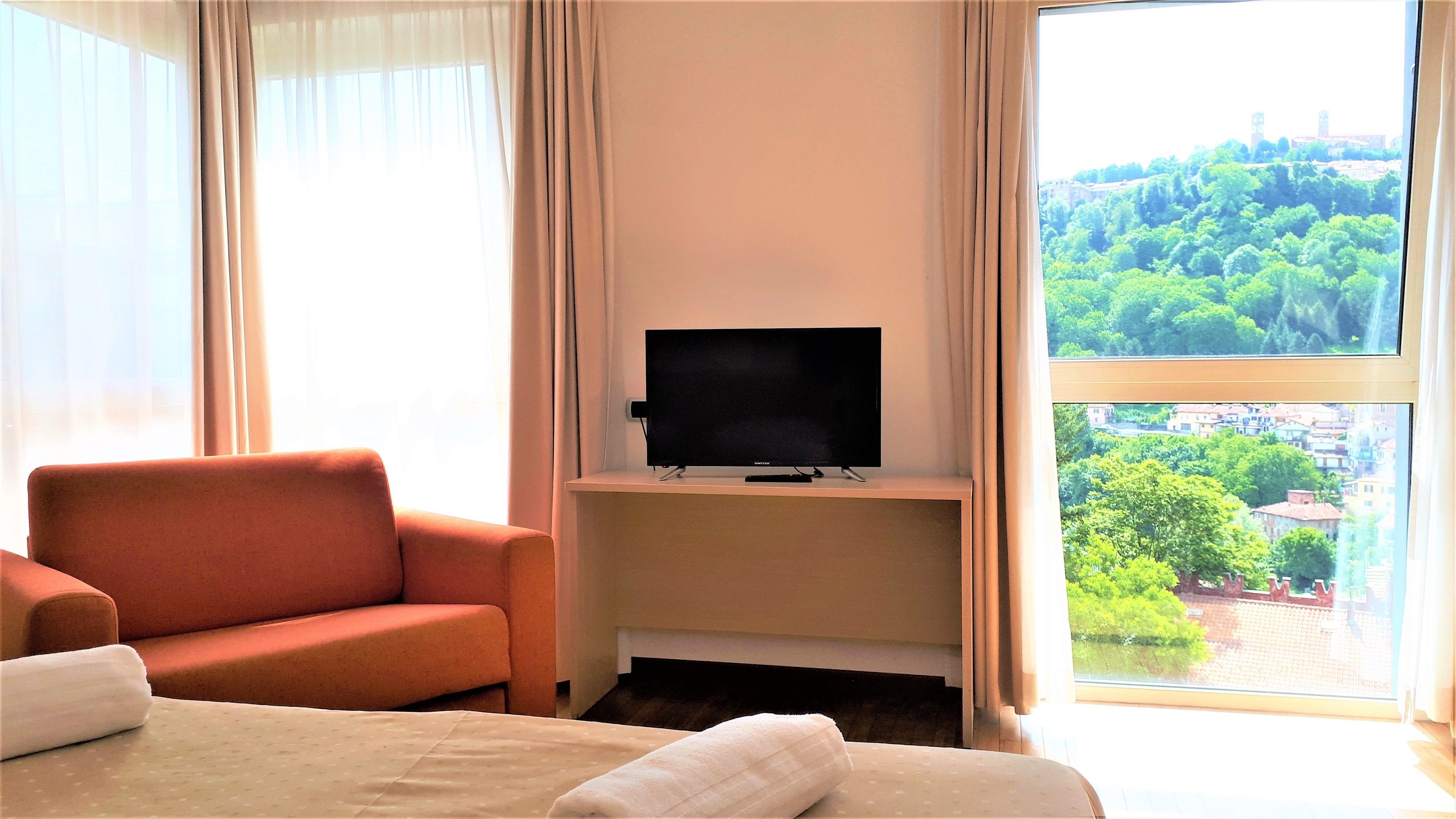 Habitación de hotel con cama, sofá, televisor sobre un soporte y un ventanal con vista a la ciudad.