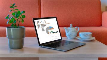 Ein Laptop, der Diagramme zeigt, steht auf einem Tisch mit Teekanne und Tassen daneben. Im Hintergrund sind ein Topf mit Pflanze und ein orangefarbenes Sofa zu sehen.