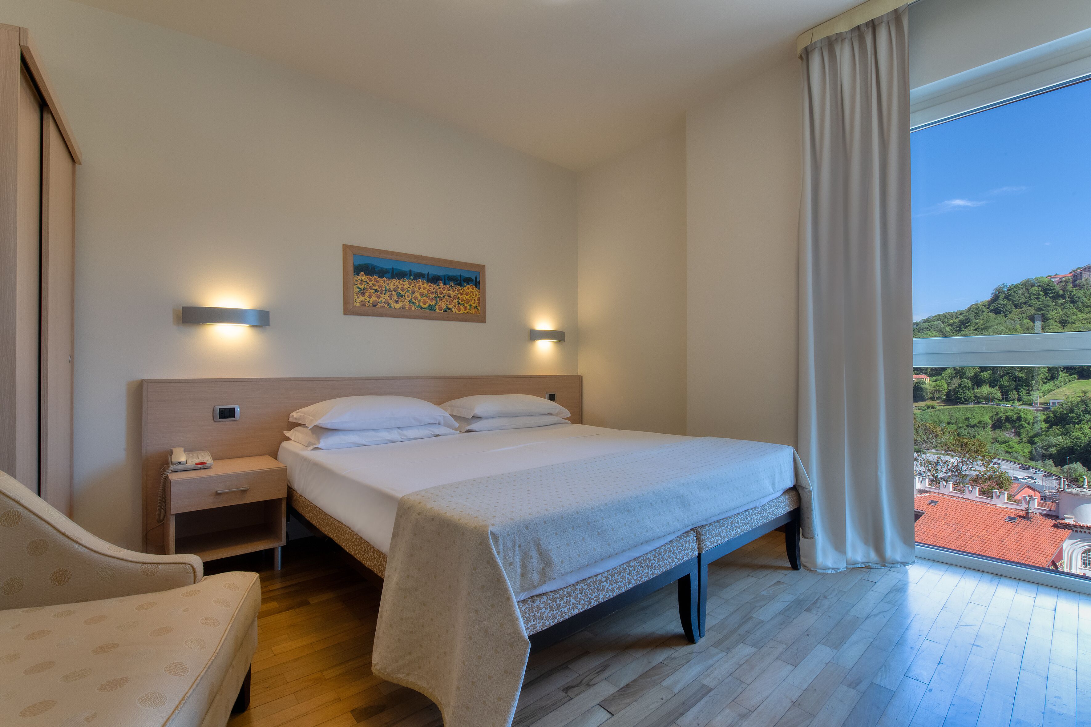 A hotel room with a bed, bedside table, lamp, and a view of a cityscape through a window.