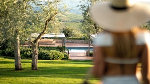 Une personne portant un chapeau blanc et un haut blanc se dirige vers un bâtiment doté d’une piscine, entouré de verdure et d’un paysage vallonné.