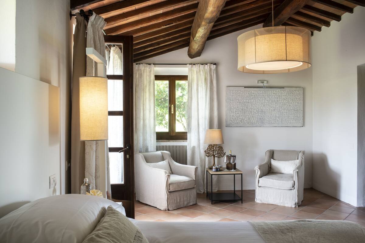 Une chambre à coucher comprenant un lit, deux fauteuils, une petite table avec une lampe, une fenêtre avec des rideaux et une œuvre d’art accrochée au mur.