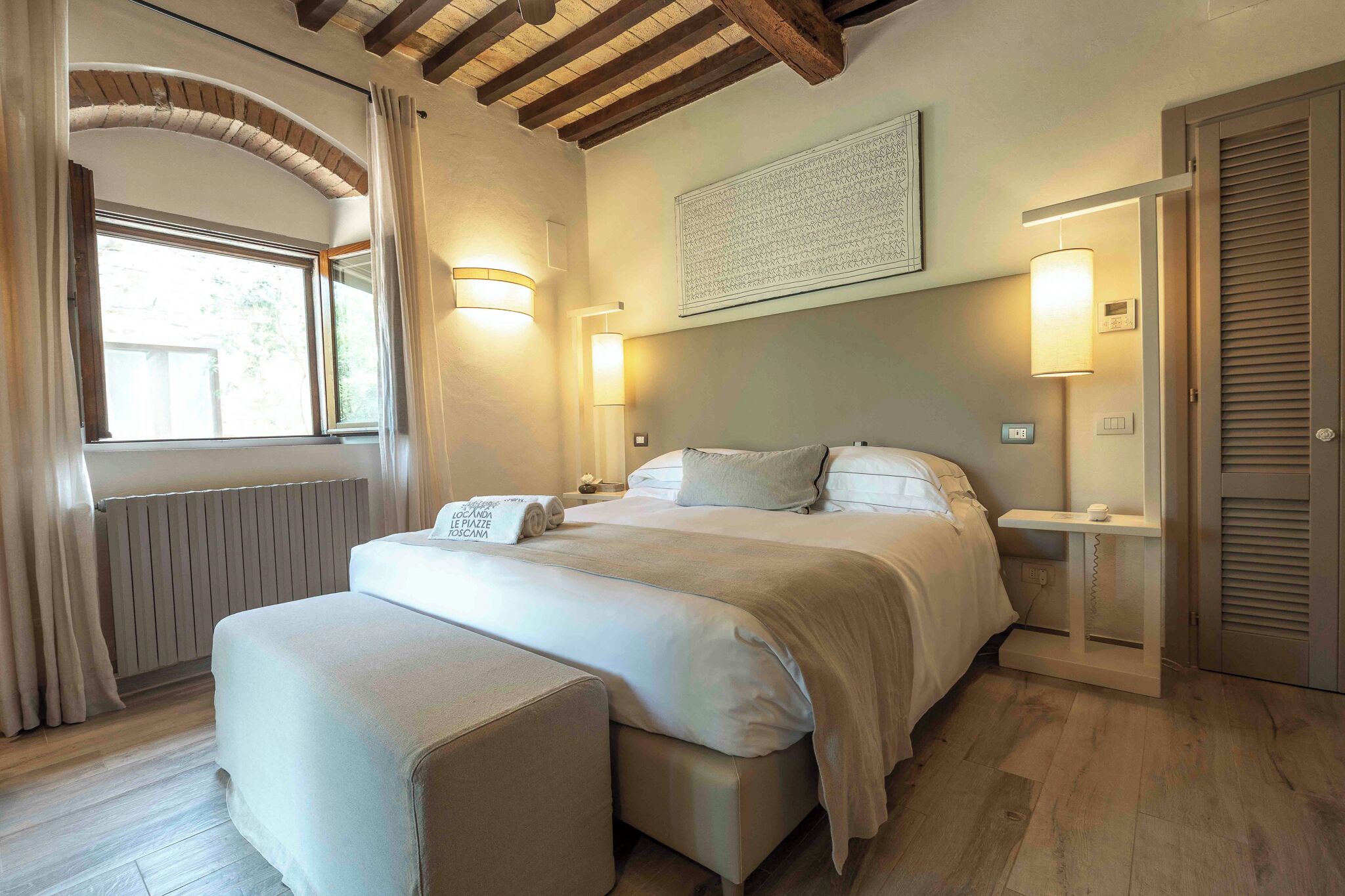 Een slaapkamer met een groot bed, een nachtkastje, een radiator, gordijnen bij een raam en een houten plafond.