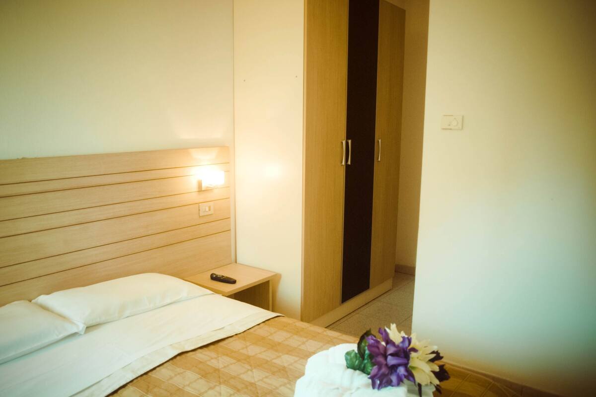 Habitación de hotel con cama, mesita de noche, control remoto y armario.