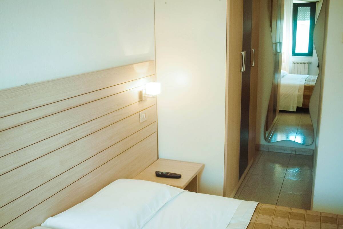 Habitación de hotel con cabecera de madera, una cama individual con ropa de cama blanca, una mesita de noche con un control remoto y vista a un baño por una puerta abierta.