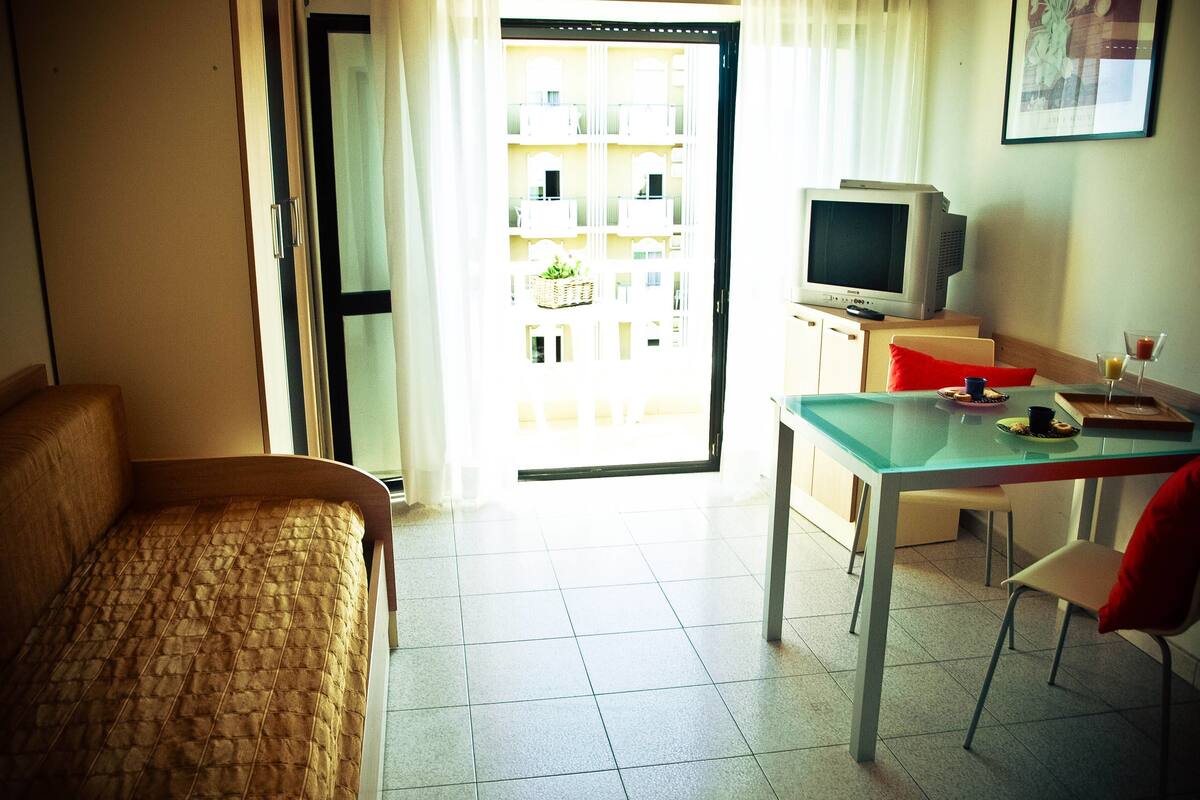 Habitación de hotel con cama, mesa de comedor con sillas, televisor y balcón con vistas a edificios.