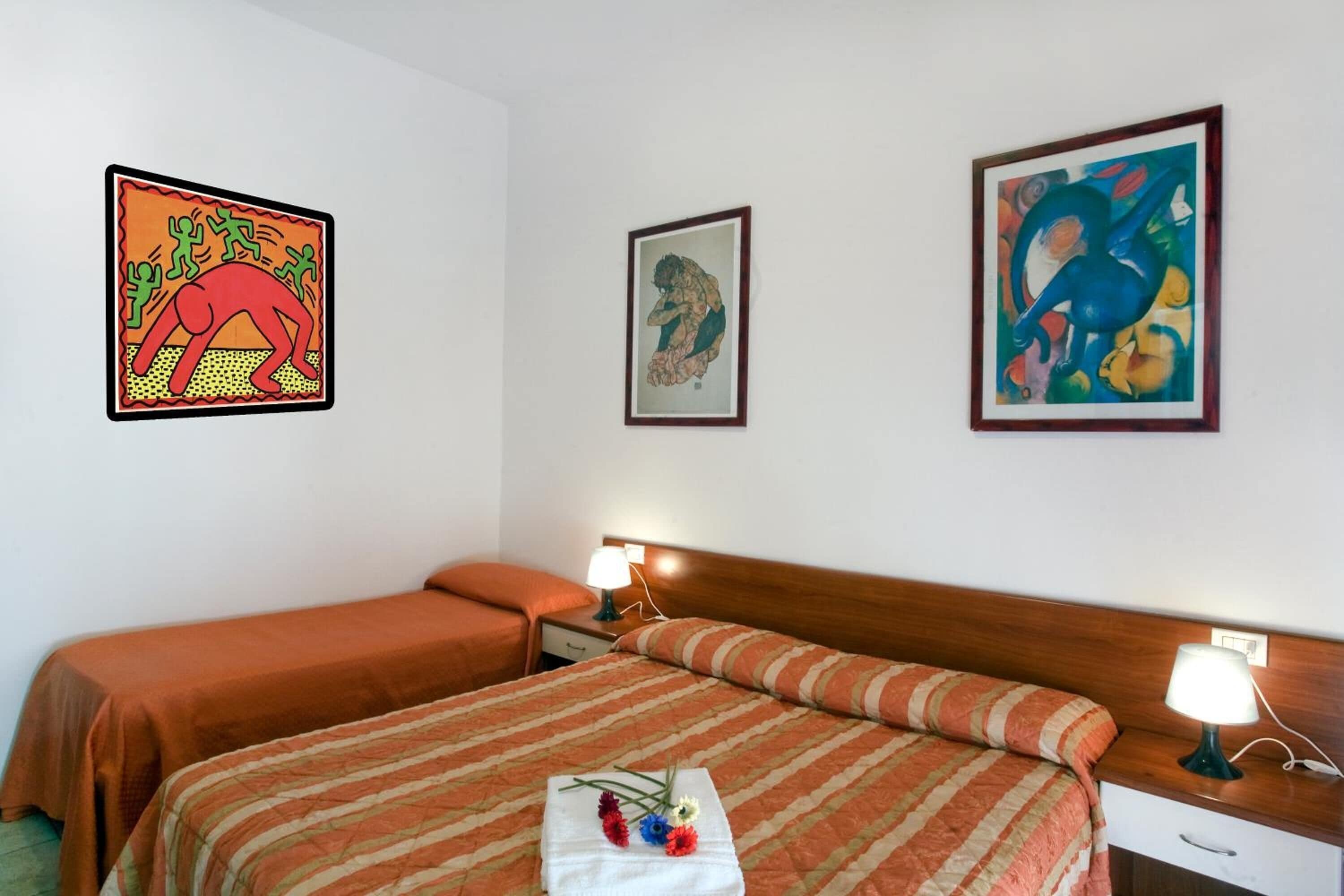 Habitación de hotel con dos camas, una mesita de noche, una lámpara y tres cuadros en la pared.