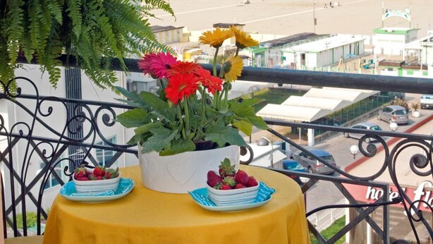 A balcony with a yellow tablecloth, a potted plant with red flowers, and a plate of strawberries. The view includes a beach and a coastal town. - Residence Auriga (Rimini)