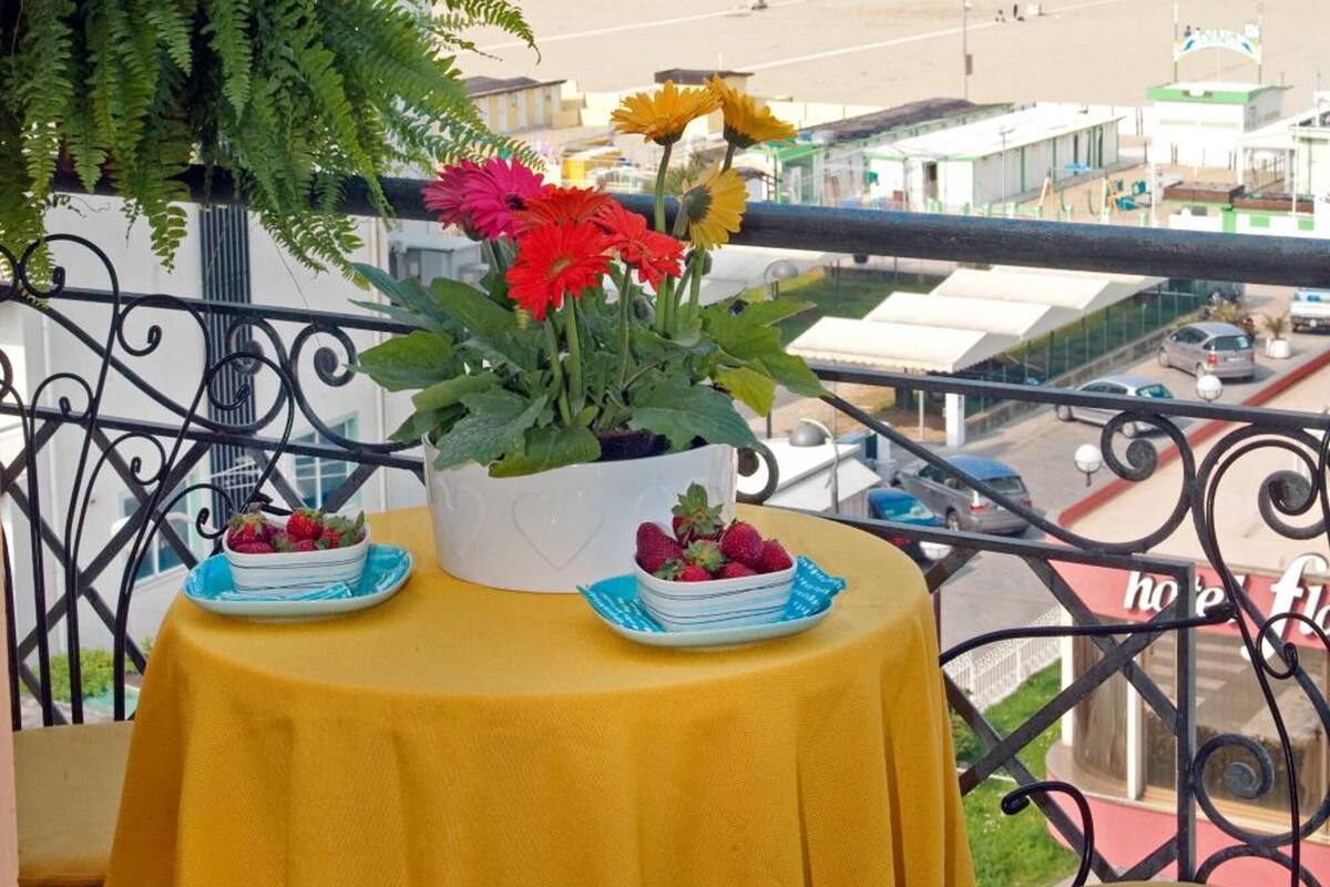 A balcony with a yellow tablecloth, a potted plant with red flowers, and a plate of strawberries. The view includes a beach and a coastal town.
