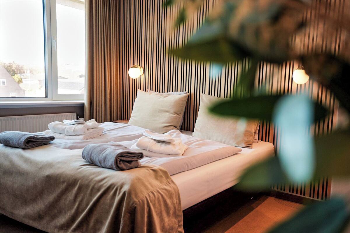 A neatly made bed with towels, a window with curtains, and a wall with vertical lines and wall-mounted lights.