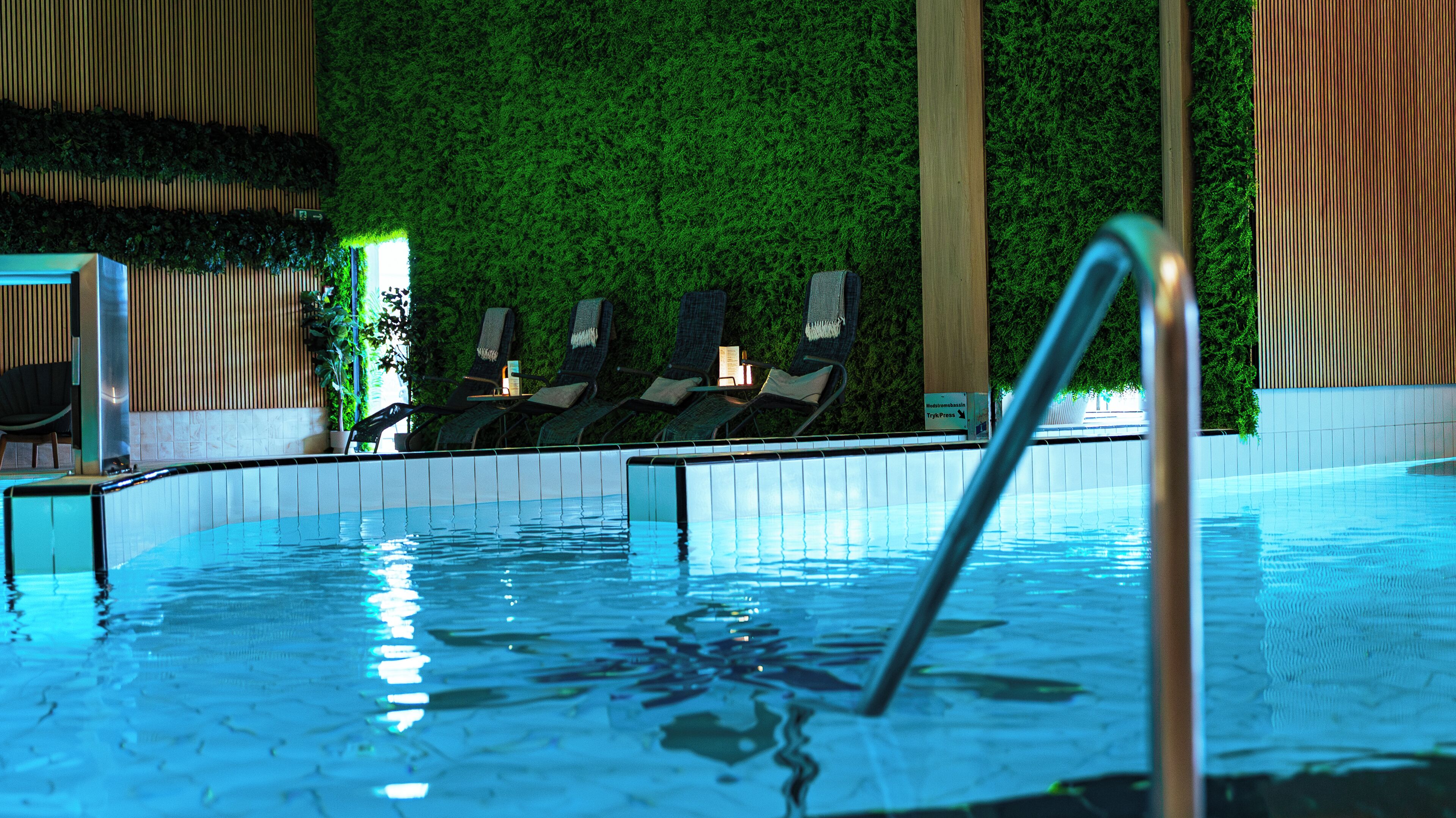 An indoor swimming pool with a clear water surface, a metal pool ladder, and lounge chairs with a green wall in the background.