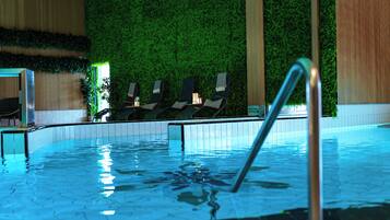 Una piscina interna con superficie d'acqua limpida, scala di metallo e sedie a sdraio, con un muro verde sullo sfondo.