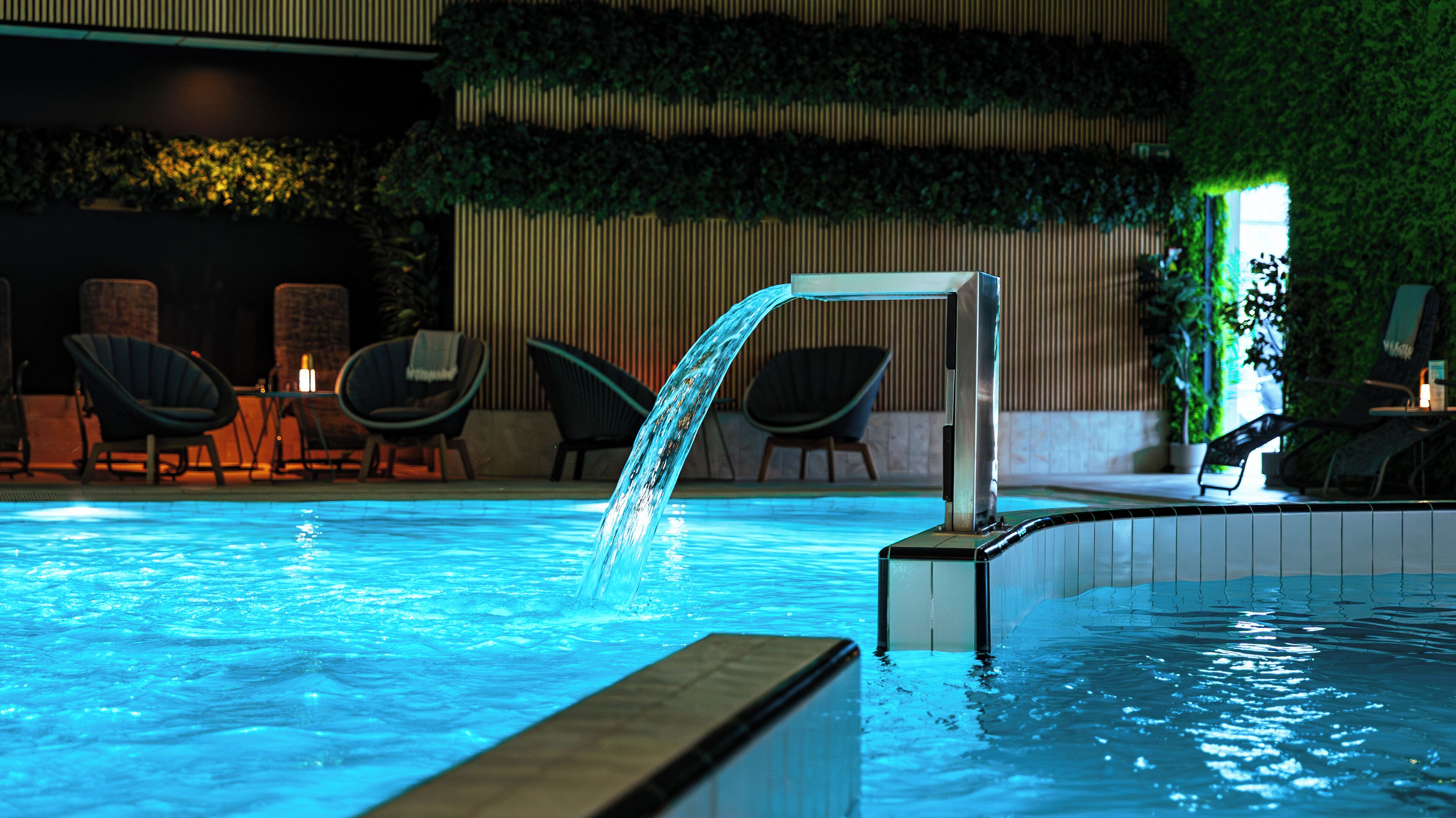 An indoor swimming pool with a waterfall feature, surrounded by lounge chairs and a wall with vertical greenery.