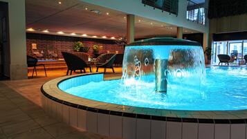 A heated indoor pool with a central water feature, surrounded by lounge chairs and a stone wall in the background.