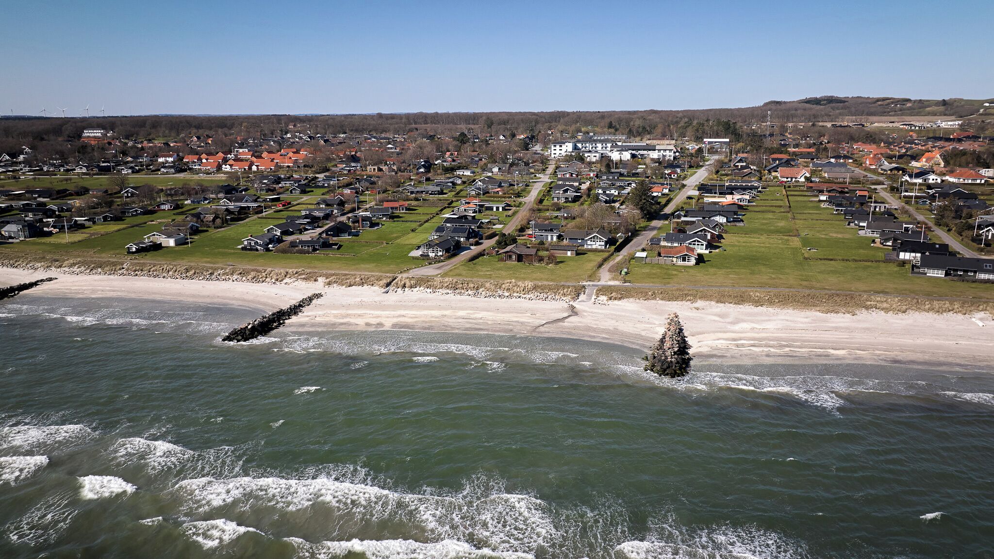An aerial view of a coastal town with houses, a beach, and wind turbines in the distance.