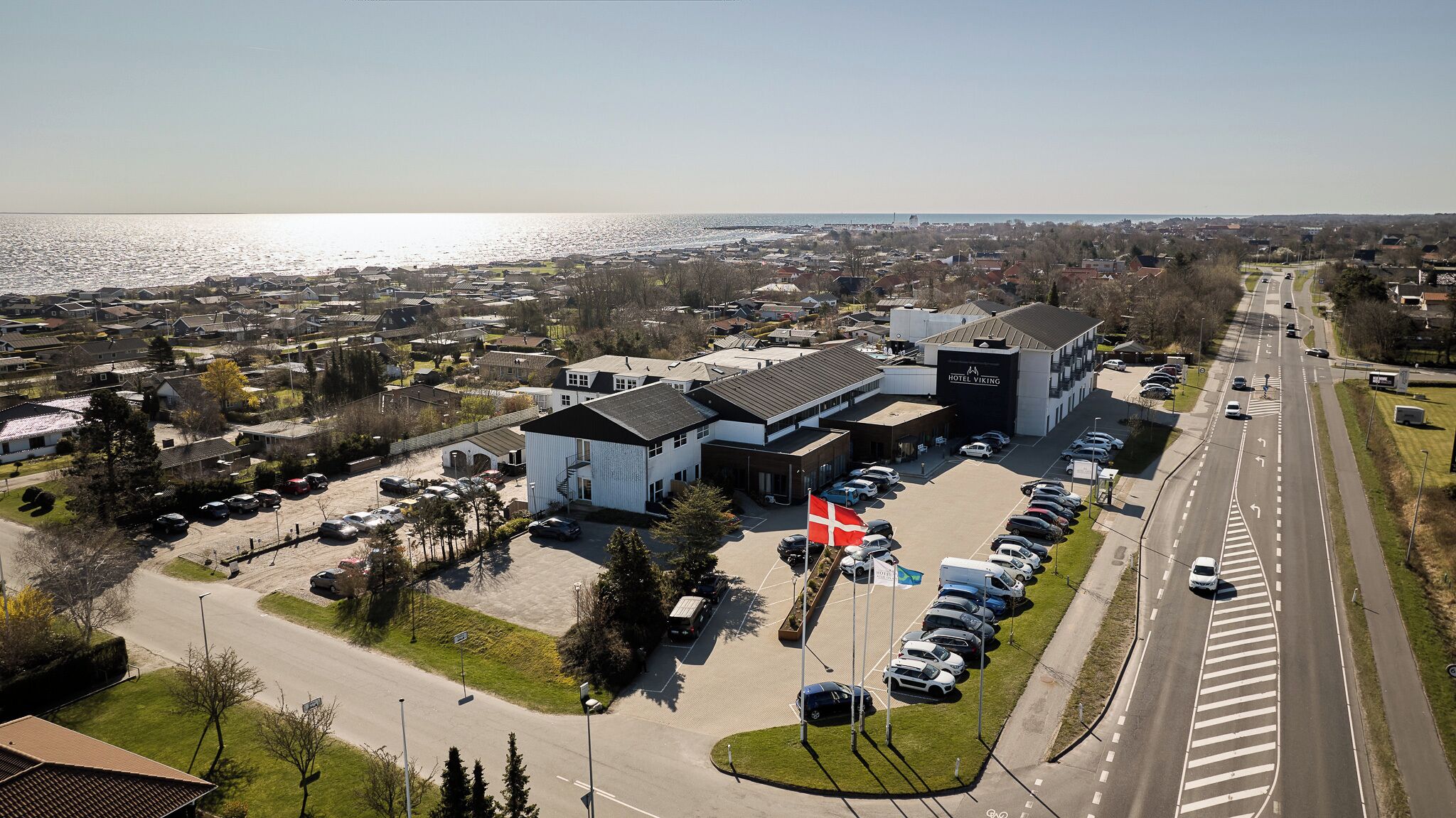A hotel with a parking lot, a road with cars, and a body of water in the background.