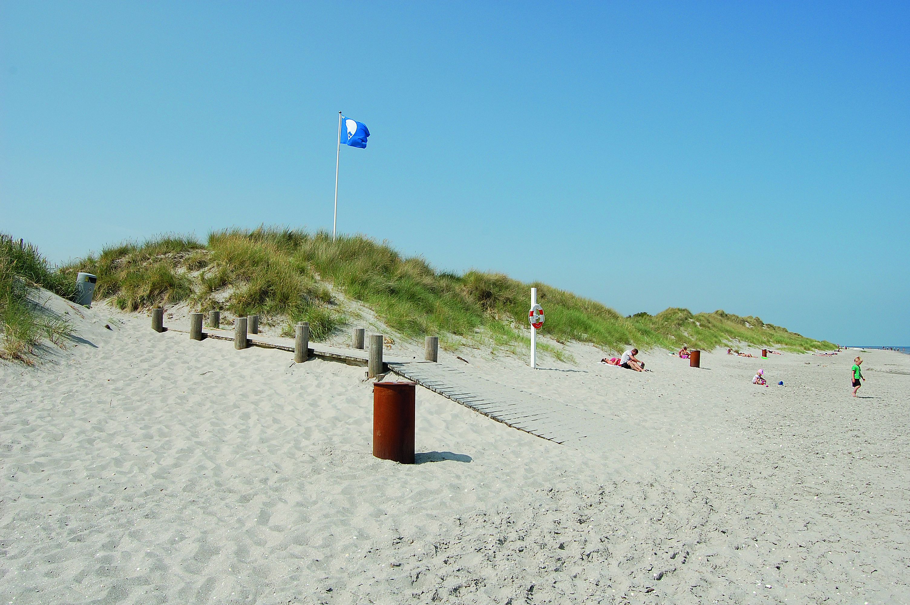 A sandy beach with a flag, wooden fence, and a trash bin.