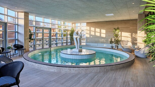 Un área de piscina interior moderna con una escultura central, rodeada de asientos y grandes ventanales.