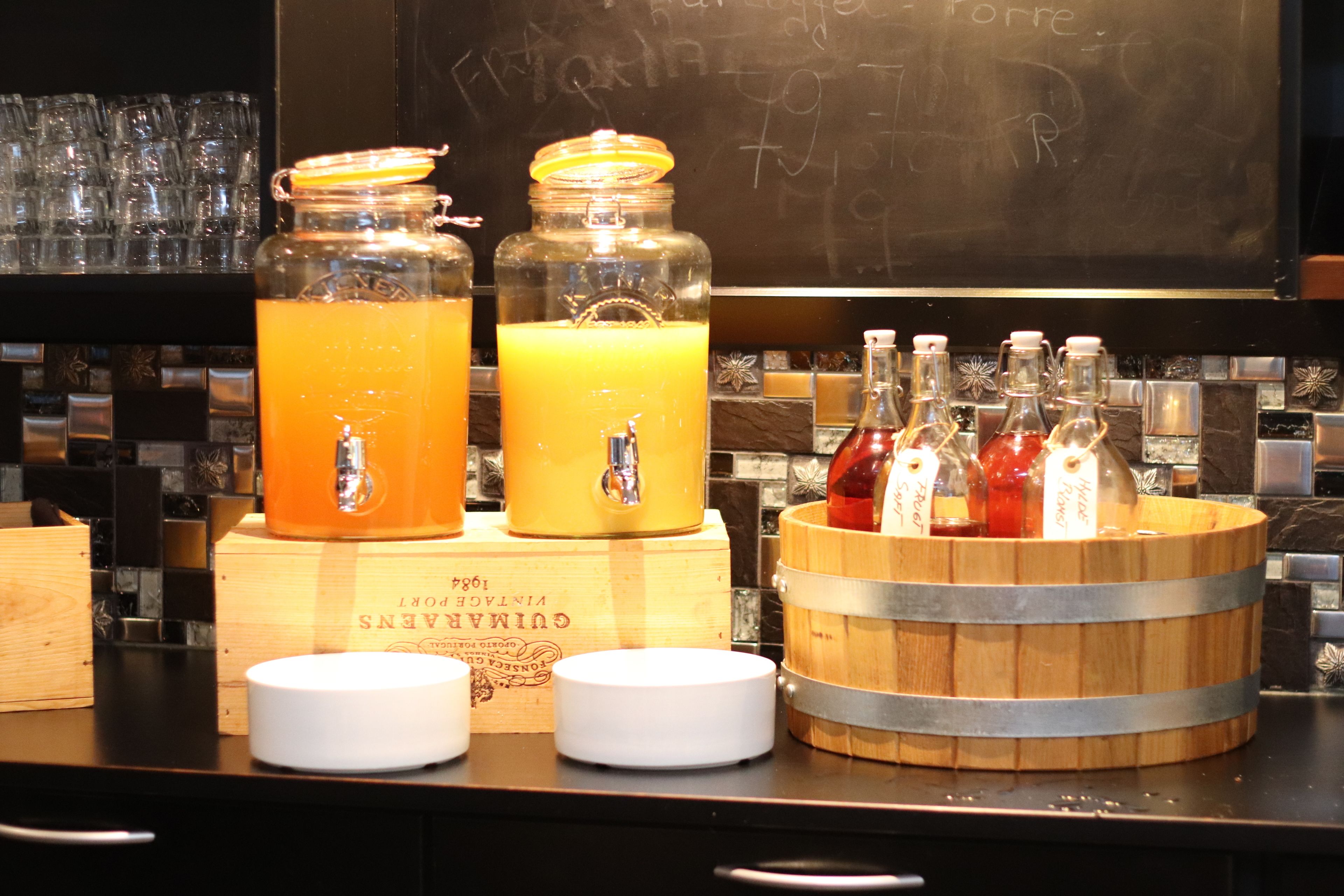 A bar area with two large glass jars of orange juice, a wooden barrel with bottles, and a chalkboard in the background.