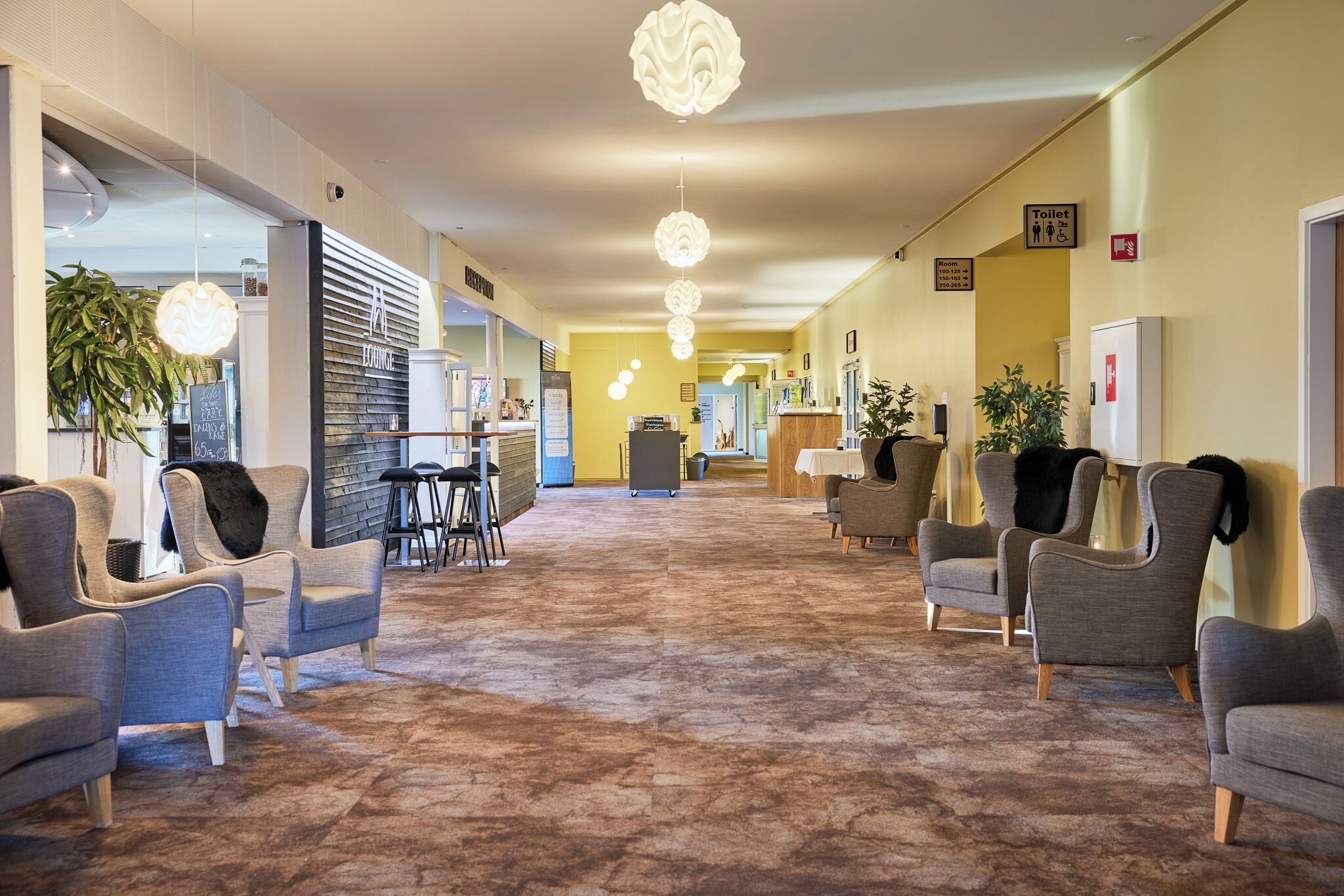 A hotel lobby with a carpeted floor, seating area with chairs, a reception desk, and hanging pendant lights.